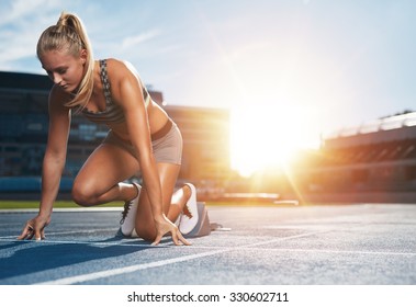 Young woman athlete at starting position ready to start a race. Female sprinter ready for sports exercise on racetrack with sun flare. - Powered by Shutterstock