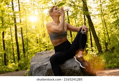 Young Woman Athlete Takes A Break At A Stone, Drinking Water, Out On A Run On A Hot Day.