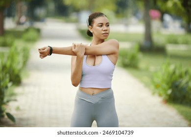 Young woman in athletic wear stretching her arm while exercising outdoors in a sunny park setting - Powered by Shutterstock