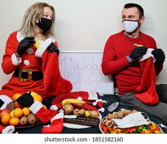 Young Woman And Attractive Man Putting Sweets And Fruits To Santa Boots For Saint Nicholas Day. Disinfection And Thermometer. Winter Holidays In 2020. Pair Wearing Face Masks To Prevent Coronavirus.