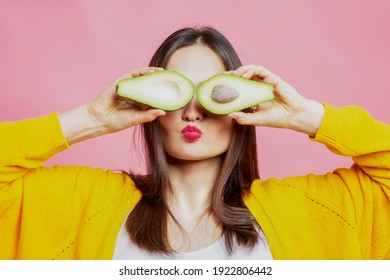 Young woman with avocado halves. Beautiful brunette in a yellow sweater. Healthy food and vegetarianism. Pink background.
