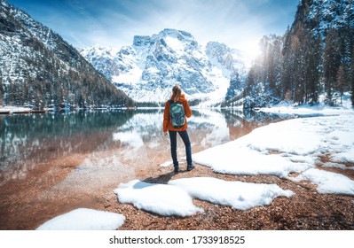 Young Woman With Backpack On The Snowy Shore Of Braies Lake With Clear Water At Sunny Bright Day In Spring. Travel. Landscape With Slim Girl, Reflection In Water, Mountains, Green Trees, Blue Sky