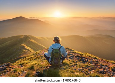 Young woman with backpack sitting on the mountain peak and beautiful mountains in fog at sunset in summer. Landscape with sporty girl, green forest, hills , sky, sunbeams. Travel and tourism. Yoga