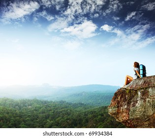 Young woman with backpack sitting on cliff and looking to a sky