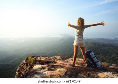 Young woman with backpack standing on cliff's edge and looking to a sky with raised hands