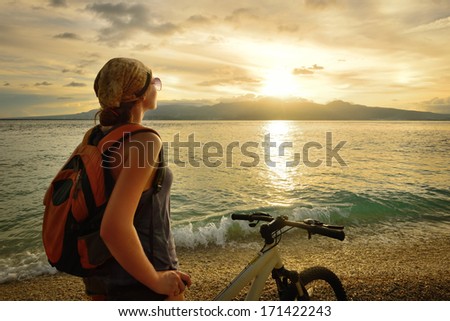 Young woman with backpack standing on the shore near his bike and enjoying the sunset over the sea on the background of the island Negros, Philippines.