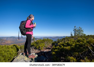 Young Woman Backpacker Reaching Her Goal Standing On Peak Of The Mountain, Holding Cup Of Coffee And Enjoying The Beautiful Landscape. Sunny Autumn Time.