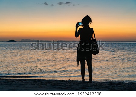 A young woman with a bag stands on the beach and photographs the sunset on a smartphone