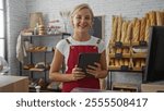 Young woman baker with short blonde hair holding a tablet in a well-lit bakery shop surrounded by bread and pastries