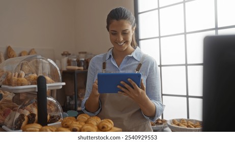 Young woman in a bakery holding a tablet, surrounded by pastries, smiling indoors, showcasing an attractive and cheerful hispanic female baker in an interior shop setting. - Powered by Shutterstock