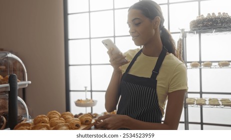 Young woman in bakery taking photo of pastries with smartphone, wearing apron, standing indoors surrounded by baked goods, shop interior in background, hispanic brunette smiling - Powered by Shutterstock