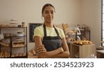 Young woman in a bakery wearing an apron with arms crossed, surrounded by pastries and bread in a cozy, well-lit shop, conveying a confident and independent business atmosphere.