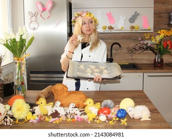 Young Woman Baking Easter Cakes In Decorated Kitchen. Celebrating Spring Holidays In 2022. Girl Wearing Chef Uniform And Flower Wreath On Head. Chicken And Rabbit Or Bunny Shapes. Big Gingerbread Lamb