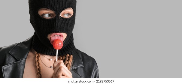 Young woman in balaclava and with lollipop on grey background with space for text