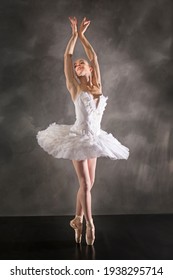 Young Woman Ballerina In White Tutu, Dancing On Pointe With Arms Overhead, In The Studio Against A Dark Background.