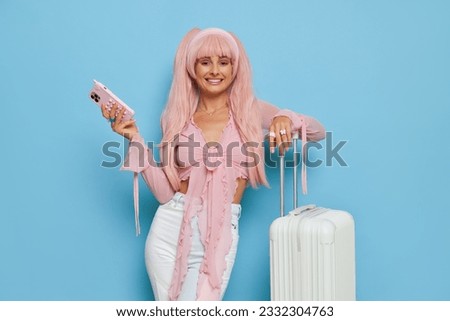 Young woman in Barbie style pink wig and pink shirt holding phone and smiling, posing on blue background with white suitcase, girl style concept, copy space