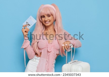 Young woman in barbie style pink shirt and hair holding passport and smiling, posing on blue background with white suitcase, girl style concept, copy space