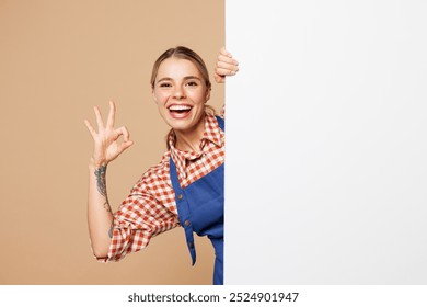 Young woman barista bartender barman wear blue apron red shirt work in coffee shop hold big white blank poster for promotional content show ok okay isolated on plain beige background. Business concept - Powered by Shutterstock