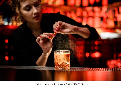 young woman bartender gently sprinkles orange peel juice on glass with cold alcoholic cocktail on the bar counter
