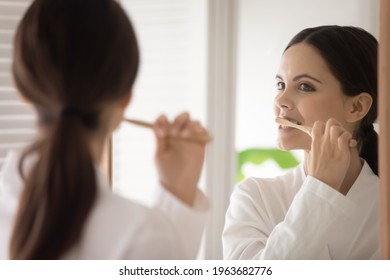Young woman in bathrobe brushing teeth with wooden bamboo toothbrush. Teen girl choosing eco friendly tools for morning bath routine, oral mouth hygiene, protection from caries. Mirror reflection