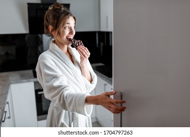 Young Woman In Bathrobe Eating Chocolate Near The Fridge, Feeling Hungry At Night. Concept Of Not Regular Eating And Overeating At Night