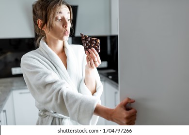 Young Woman In Bathrobe Eating Chocolate Near The Fridge, Feeling Hungry At Night. Concept Of Not Regular Eating And Overeating At Night