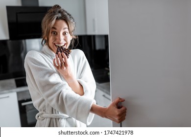 Young Woman In Bathrobe Eating Chocolate Near The Fridge, Feeling Hungry At Night. Concept Of Not Regular Eating And Overeating At Night