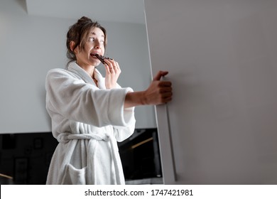 Young Woman In Bathrobe Eating Chocolate Near The Fridge, Feeling Hungry At Night. Concept Of Not Regular Eating And Overeating At Night