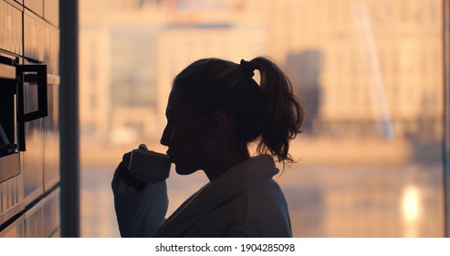 Young Woman In Bathrobe Making Coffee With Coffee Machine In Kitchen At Home And Enjoying Sunrise View In Panoramic Window. Side View Of Happy Female Enjoying Coffee In Morning