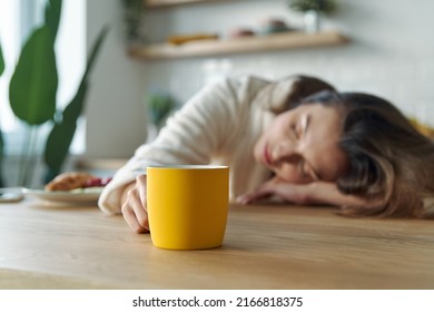 Young woman in bathrobe napping with cup of coffee while sitting at the kitchen counter at home