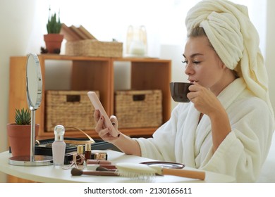 Young woman in bathrobe sitting at vanity, drinking coffee and reading news on smartphone - Powered by Shutterstock