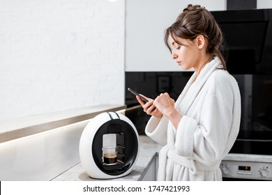 Young Woman In Bathrobe Using A Smart Phone While Making Coffee With A Coffee Machine On The Kitchen At Home