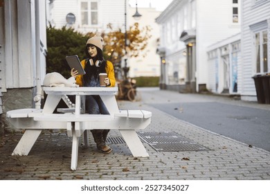 A young woman in a beanie and headphones sits at an outdoor table with coffee, browsing on her tablet in a quiet town street. - Powered by Shutterstock