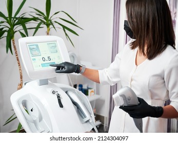 Young Woman Beautician In Black Gloves Using Device In Modern Clinic. Female Doctor Cosmetologist In White Lab Coat Pressing Start On Display Of Ultrasound SMAS Lifting Machine.