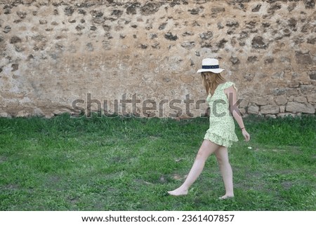 Young woman, beautiful and blonde in a yellow dress and hat, walking barefoot on the green grass, calm and relaxed. Spring concept, feet, grass, fields, meadows.