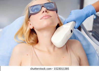 Young Woman At Beauty Clinic Cosmetology Service Lying On Medical Bed Wearing Safety Goggles Looking Up Pensive While Female Doctor Using Photorejuvenation Laser Device On Her Neck Skin