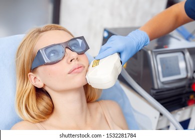 Young Woman At Beauty Clinic Cosmetology Service Lying On Medical Bed Wearing Safety Goggles Looking Up Thoughtful While Female Doctor Using Photorejuvenation Laser Device On Her Face Close-up