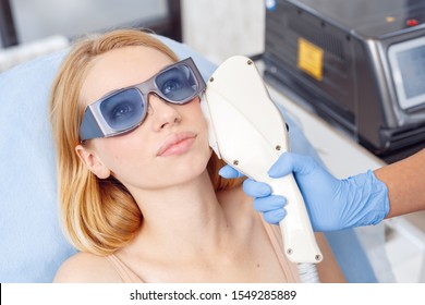 Young Woman At Beauty Clinic Cosmetology Service Lying On Medical Bed Wearing Safety Goggles Looking Up Pensive While Female Doctor Using Photorejuvenation Laser Device On Her Face Close-up