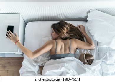 Young Woman In Bed Just Waking Up Tries To Turn Off Her Cell Phone Alarm. Concept Of New Technologies And Home.