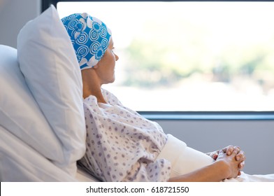 Young woman in bed suffering from cancer. Thoughtful woman battling with tumor looking out of window. Young patient with blue headscarf recovery in hospital on bed.
