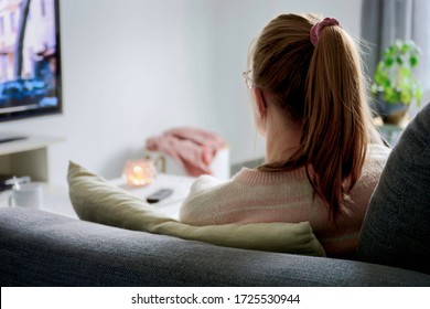 A Young Woman From Behind With Ponytail And Glasses Watching TV While Sitting On Sofa In Living Room At Home. Woman Binge Watching Tv Via Online Streaming Platform