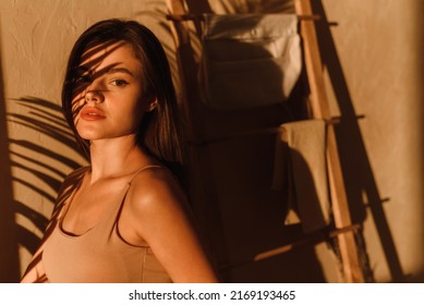A young woman in a beige bodysuit enjoys the sunbeams in a modern bathroom. Shadows from tropical leaves. - Shutterstock ID 2169193465