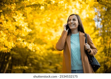 Young woman in beige coat and light blue sweater is happily talking on her smartphone while walking in a sunlit autumn park. The bright yellow foliage and warm light create a vibrant, cheerful scene - Powered by Shutterstock