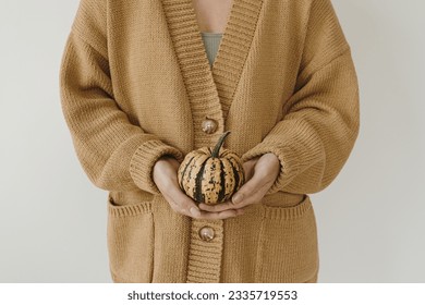 Young woman in beige knitted cardigan hold in hand decorative pumpkin over white wall