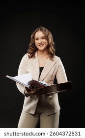 Young woman in beige suit holding an open file folder, standing confidently against a dark background. Studio lighting, professional pose, formal attire