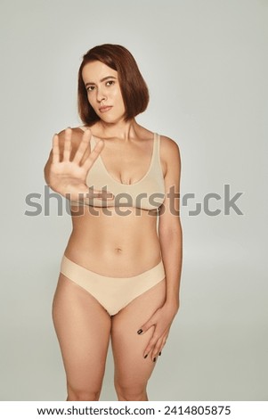 young woman in beige underwear gesturing and showing stop on grey background, body shaming