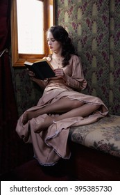 Young Woman In Beige Vintage Dress Of Early 20th Century Reading Book In Coupe Of Retro Railway Train