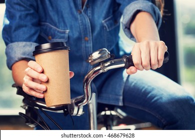Young woman with a bike holds disposable brown paper cup of coffee. Copy space on cup.