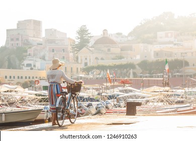 Young Woman With Bike At Ponza Island Harbor Pier In Italy. Tourist With Large Hat, Fashion Shirt And Colorful Skirt. Basket With Wine And Flowers In Front Of Shops And Boats.