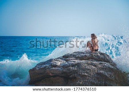 Young woman in bikin on a beach by the sea, a wave is breaking on a stone.